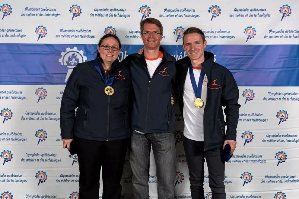 Deux étudiants médaillés et un enseignant devant le mur indiquant : Olympiades québécoises des métiers et des technologies