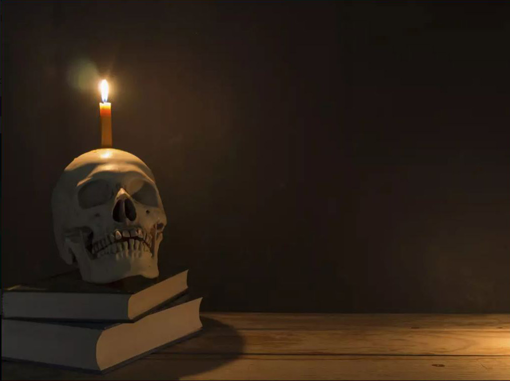 Une bougie allumée repose sur un crâne humain. Celui-ci est déposé sur une pile de livres.
