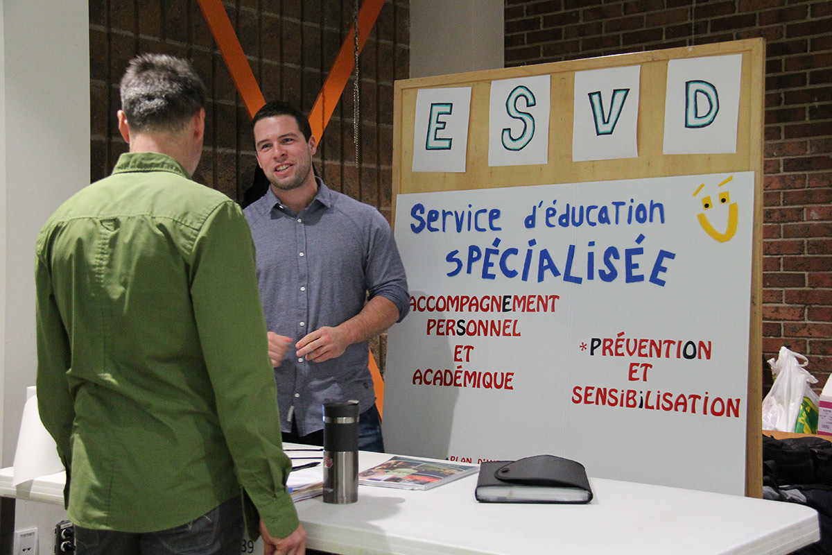 Étudiant accueillant un visiteur à son kiosque sont l'affiche indique : ESVD Service d'éducation spécialisée, accompagnement personnel et académique, prévention et sensibilisation