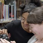 Deux étudiantes s'entraident avec la manipulation d'une caméra photo, dans la bibliothèque
