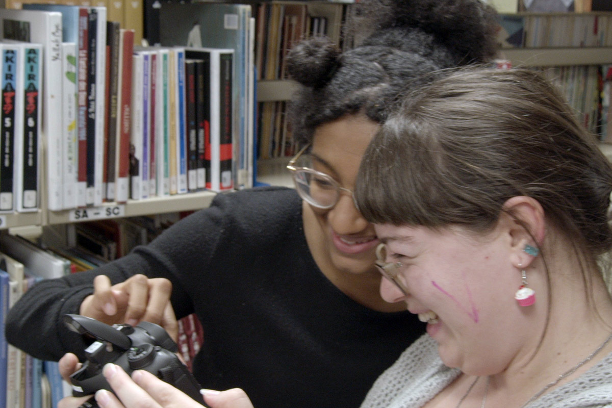 Deux étudiantes s'entraident avec la manipulation d'une caméra photo, dans la bibliothèque