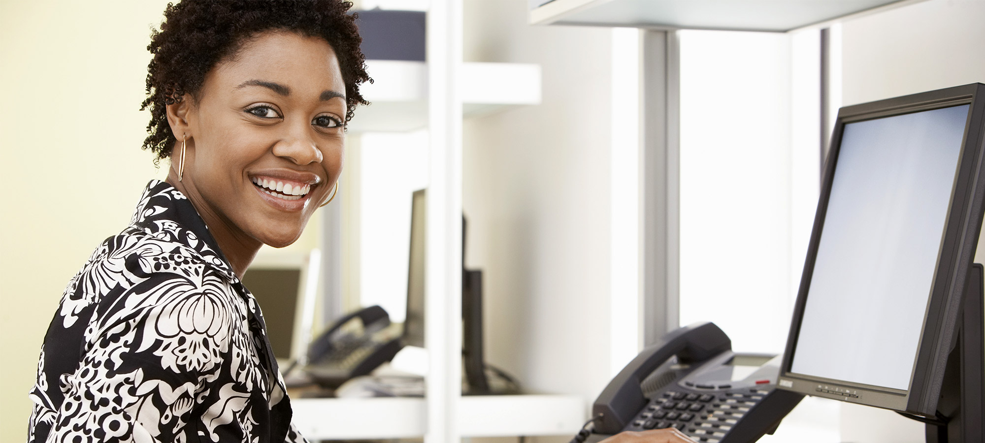 Femme souriante assise à un poste de travail informatique dans un bureau vitré
