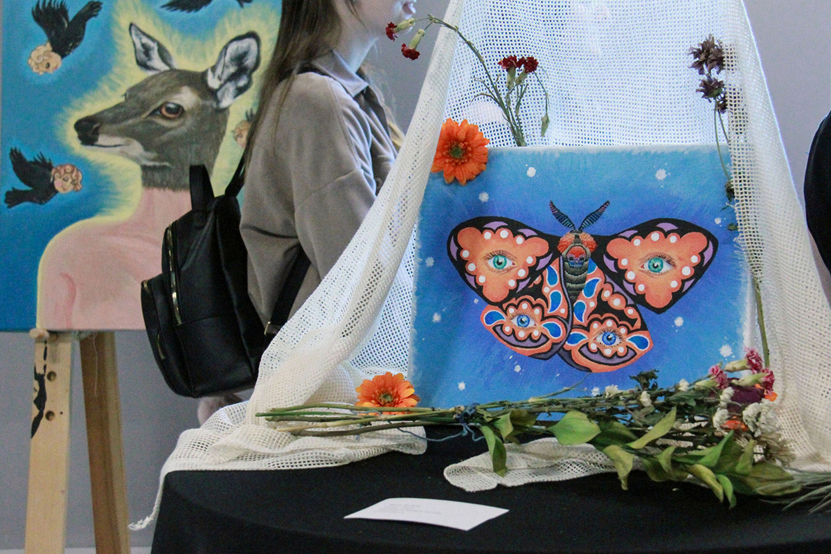En premier-plan toile avec un papillon peint, en arrièrecplan jeune fille de côté et toile avec chevreuil