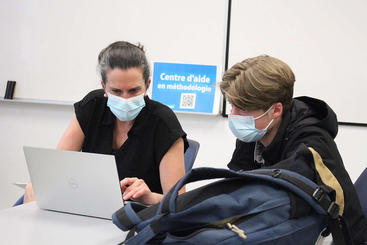 Une enseignante aide un étudiant devant un ordinateur portable, en arrière-plan l'affiche Centre d'aide en méthodologie