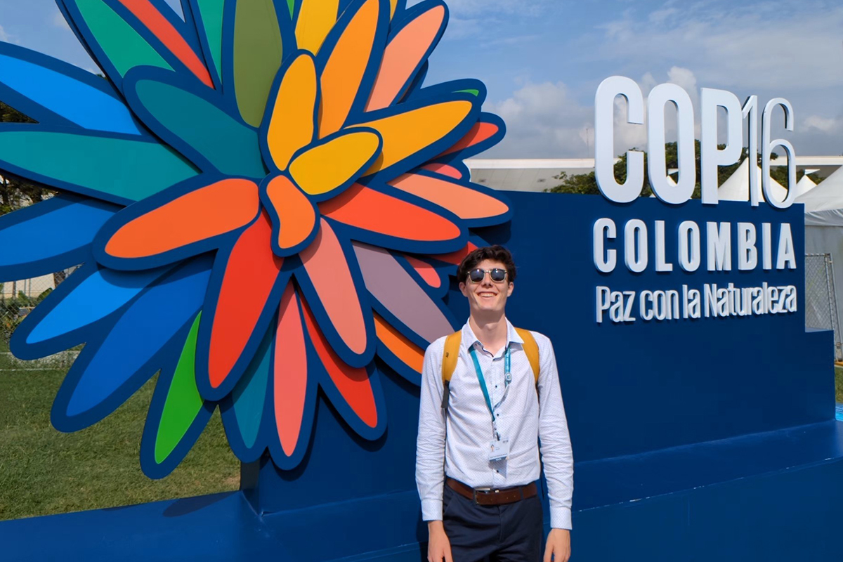 Un jeune homme debout devant un panneau de la COP16 où il y a une grosse fleur